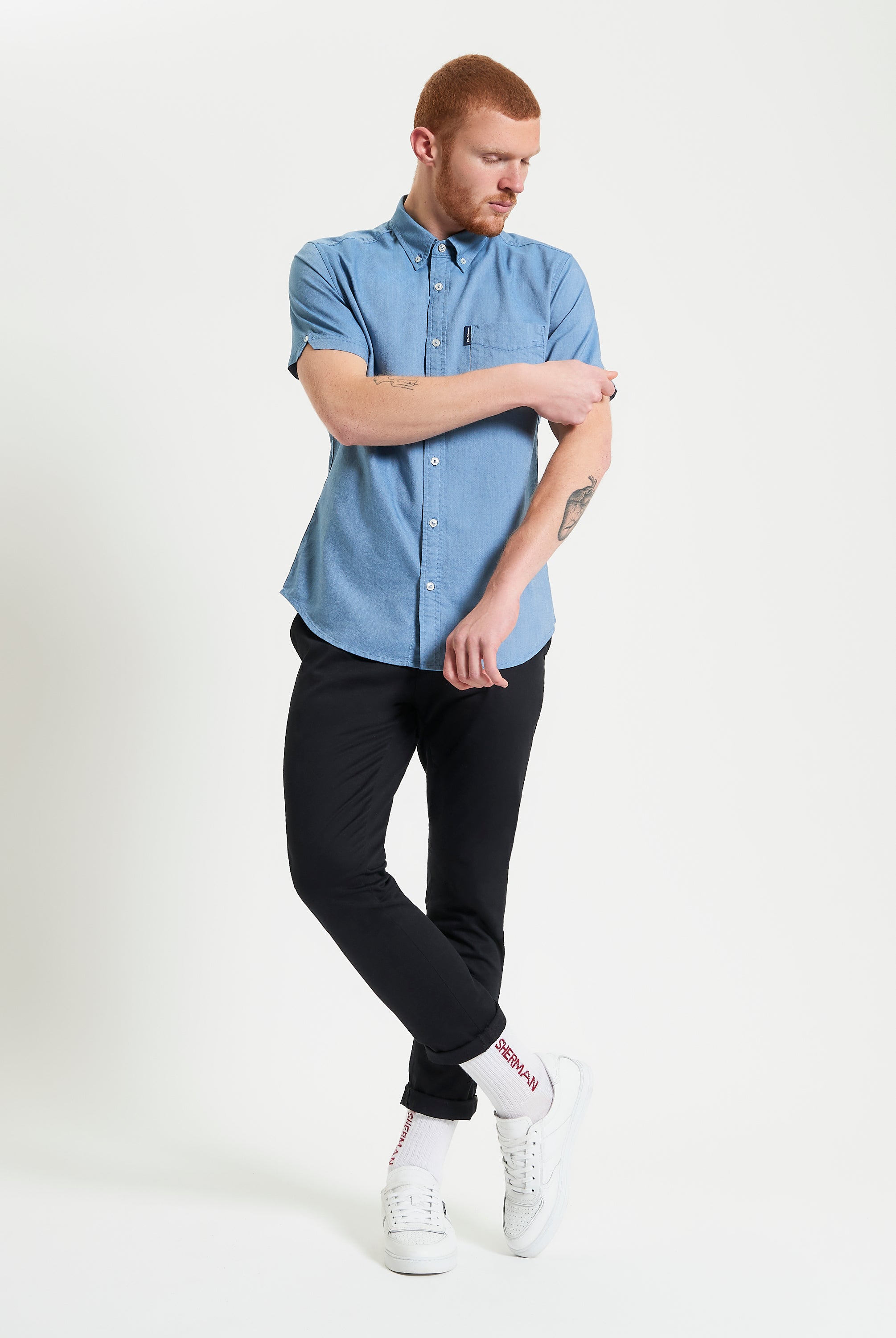 Model wearing a blue Signature Oxford Short Sleeve Shirt with black pants and white sneakers, showcasing a modern style.