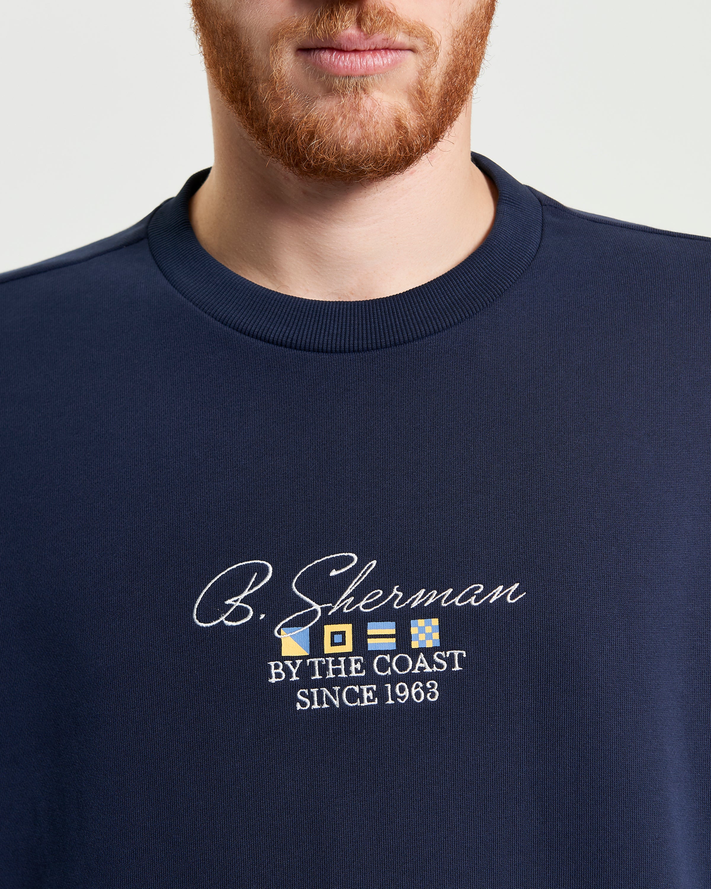 Close-up of a man wearing a navy sweatshirt featuring B. Sherman embroidery and the phrase BY THE COAST since 1963.