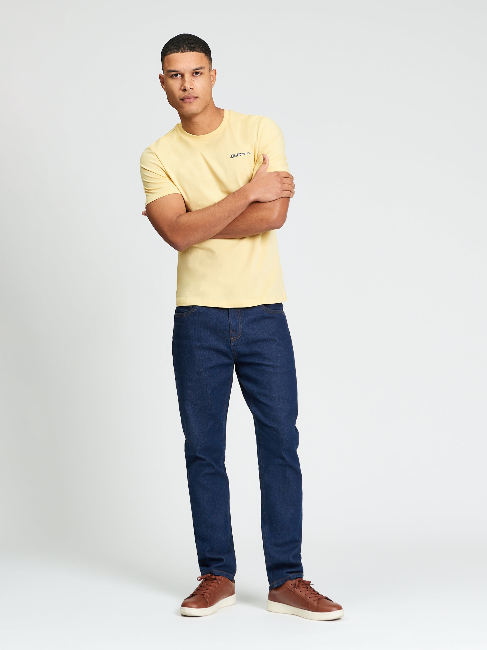 Signature Tee – Yellow crew neck t-shirt with a branded silicon badge, paired with dark jeans.